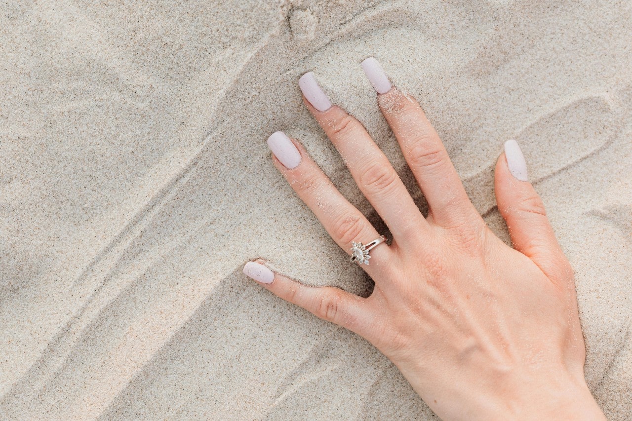 a woman’s hand in the sand, wearing a white gold engagement ring