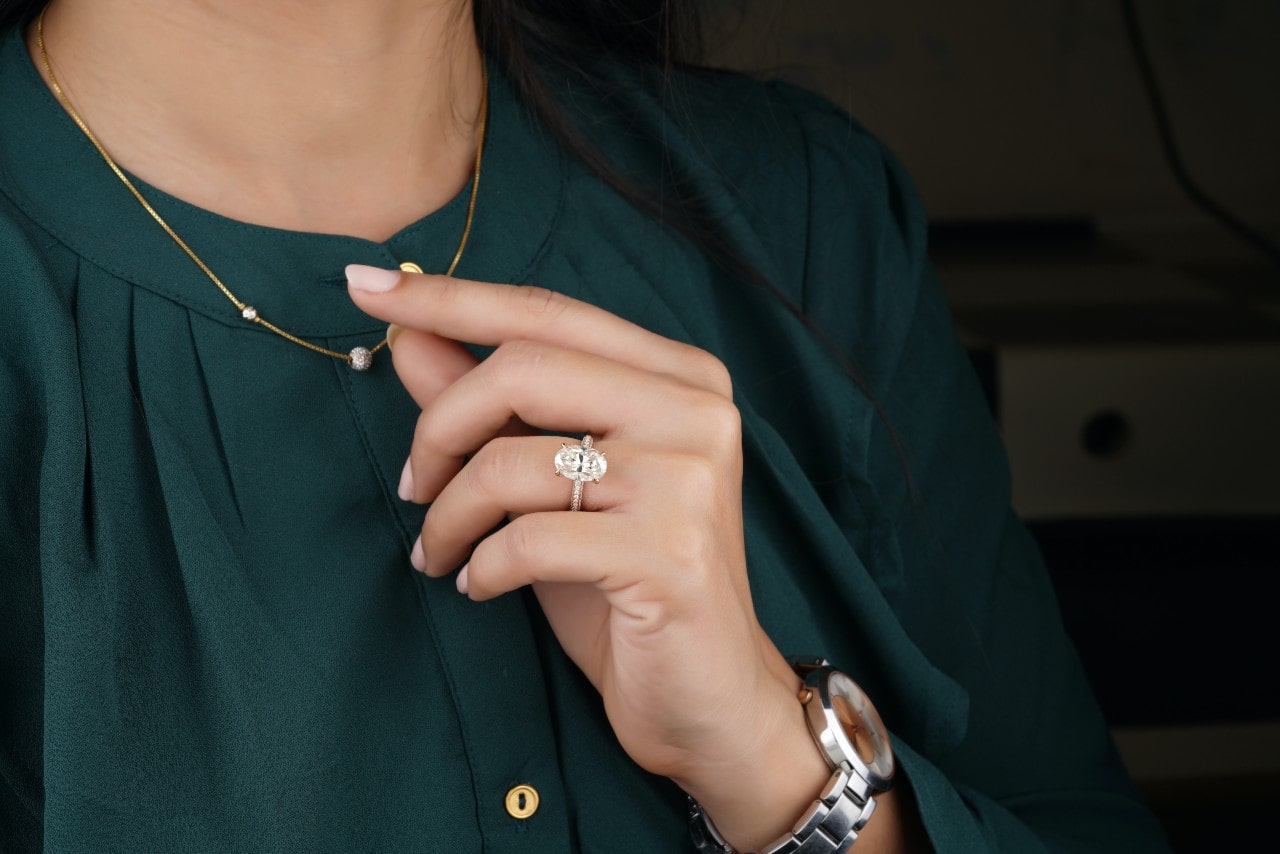 a woman wearing an oval cut engagement ring touches a necklace on her blouse.