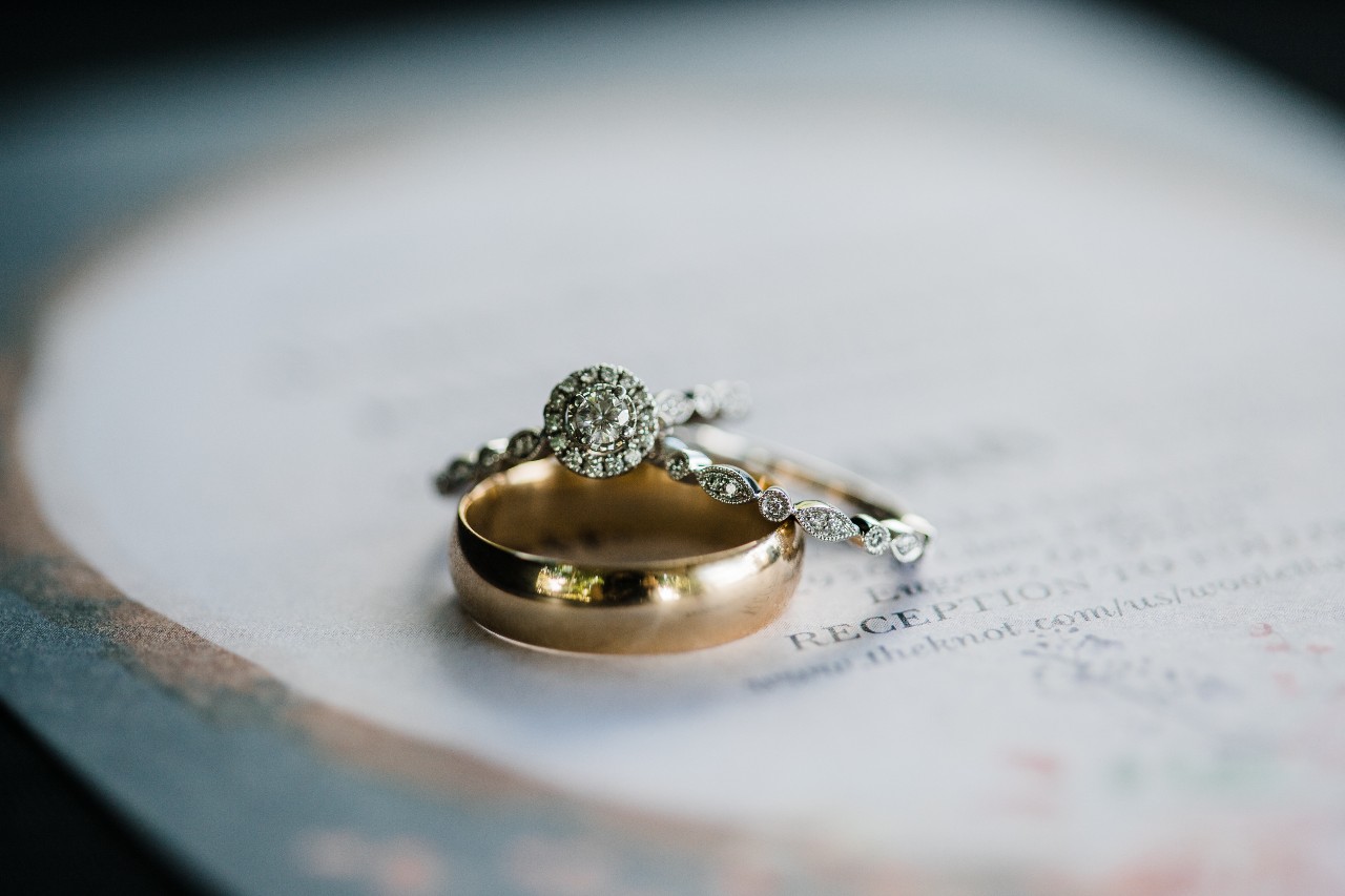 A vintage halo engagement ring with a matching band and gold band sit on a wedding invitation.