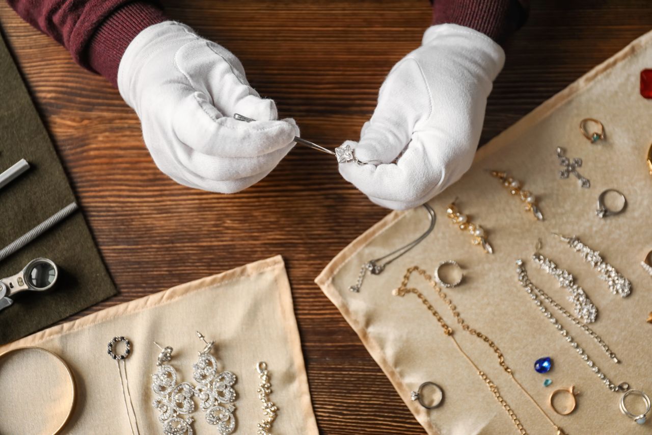 A jeweller wearing white gloves carefully inspecting a diamond necklace with precision tools surrounded by various jewelry pieces on cloth displays.