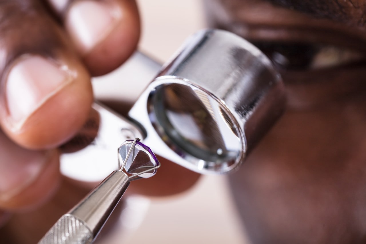 A close-up of a jeweler examining a faceted gemstone under a magnifying loupe, held with metal tweezers.