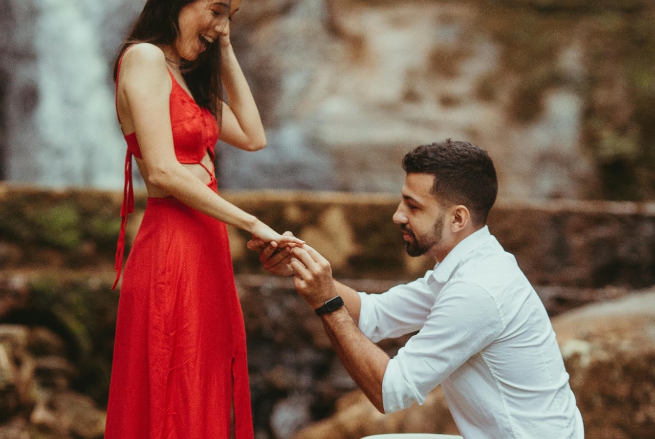Man kneeling to propose, holding a woman's hand as she smiles in a red dress, with a blurred waterfall in the background.