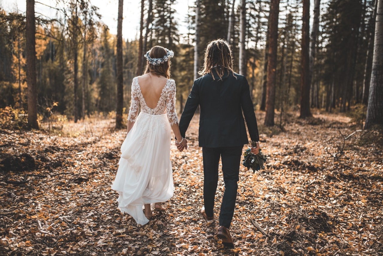 A bride in a backless lace wedding dress and a groom in a dark grey suit walk in the autumn forest, covered in fallen leaves.
