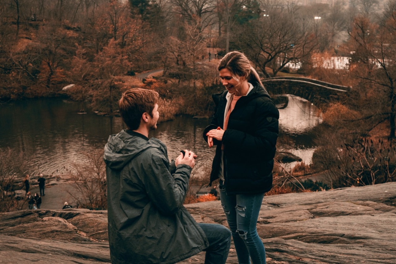 A man in a grey jacket proposing to a woman in ripped jeans in front of a fall park scenery.