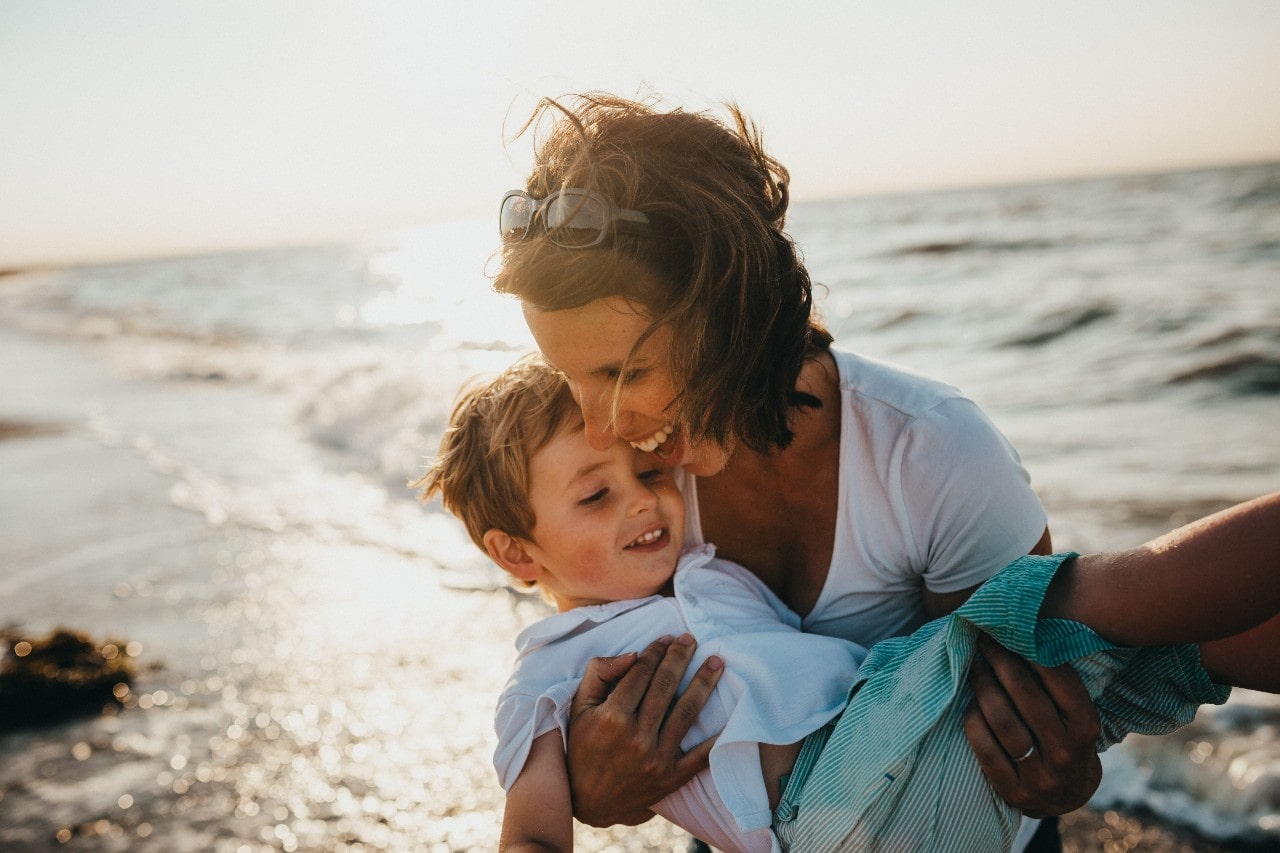 A view of a mother holding her son as they happily play at the beach in front of gently rolling waves.