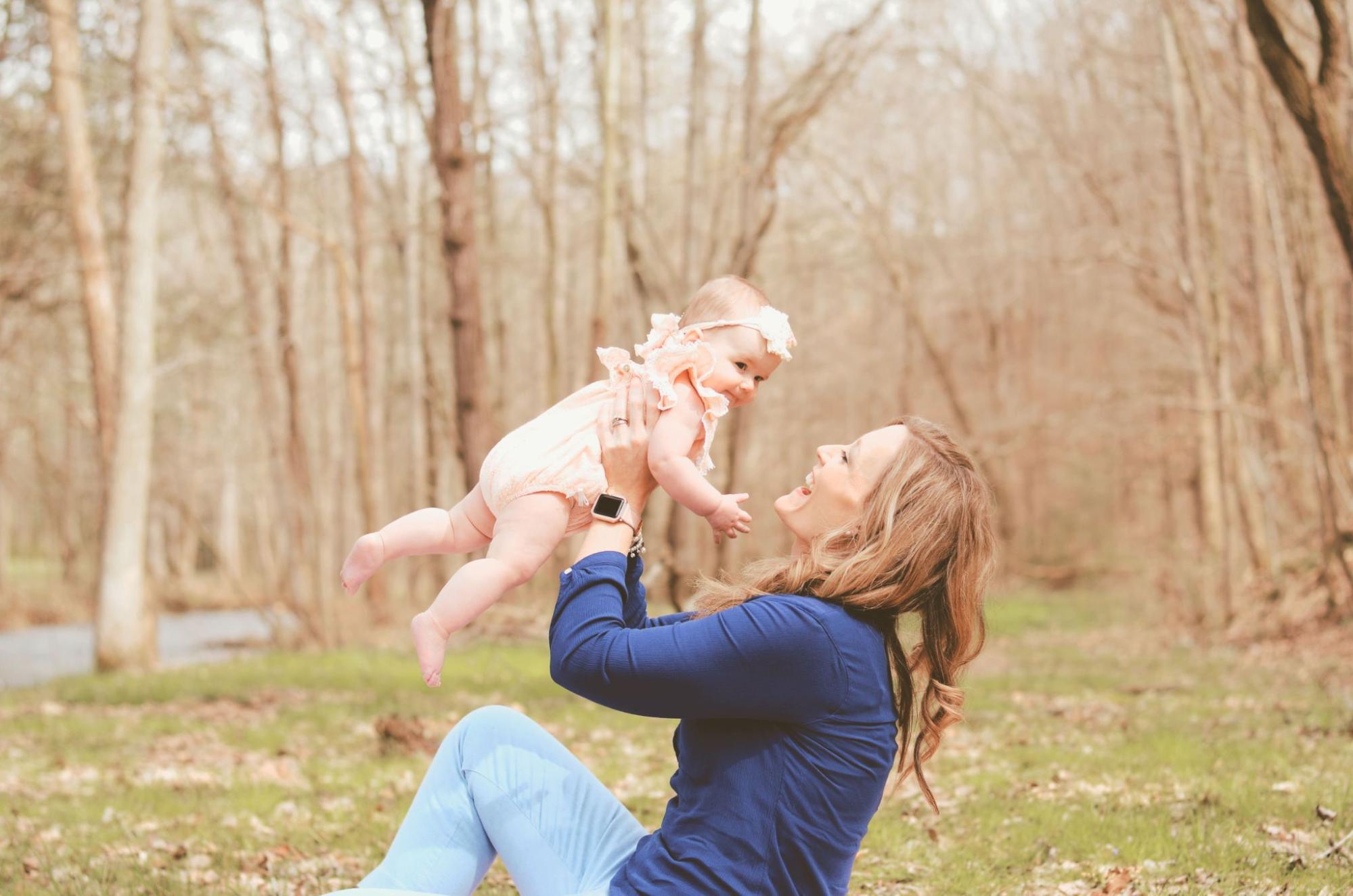 A mother in a deep blue long sleeve holds her infant in front of a forestry scenery.