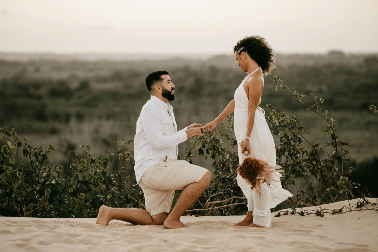 A man in white shirt and shorts kneels on a sand, proposing to a woman in a white dress.
