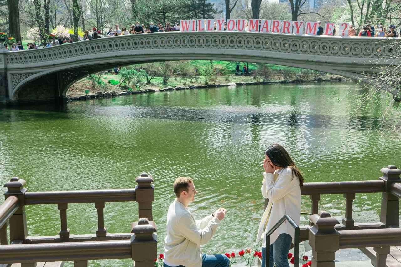 A man kneels in front of the river and bridge, proposing to a woman in white shirt and jeans.