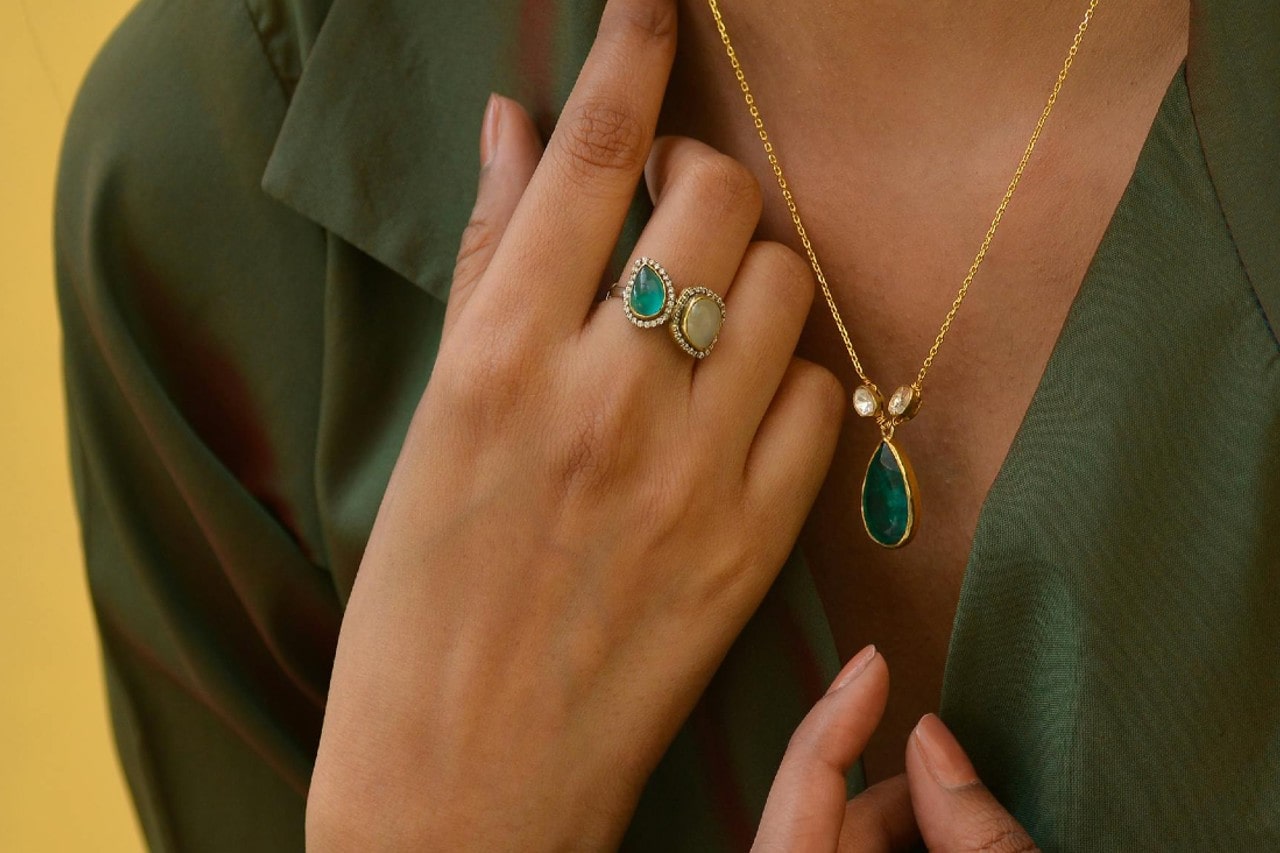A person wearing a green blouse displays elegant jewelry: a gold necklace with a teardrop pendant and a dual-stone ring.