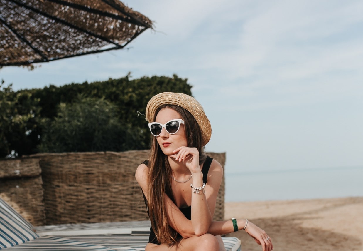 A woman in a black swimsuit and straw hat relaxes on a beach chair under an umbrella, wearing white sunglasses and charm bracelet