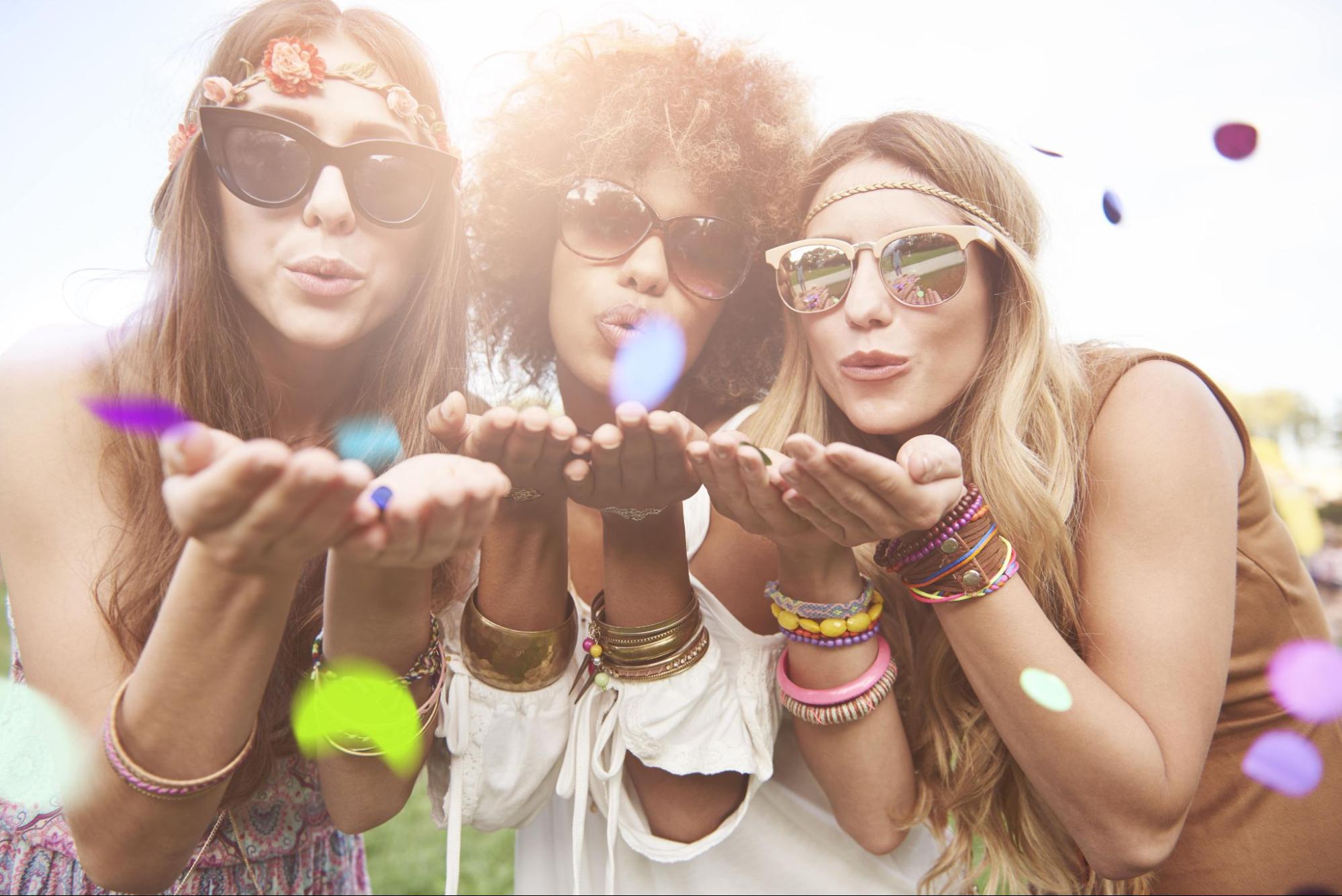 Three women dressed in boho-chic attire blow confetti toward the camera, wearing colourful accessories and sunglasses at an outdoor festival.