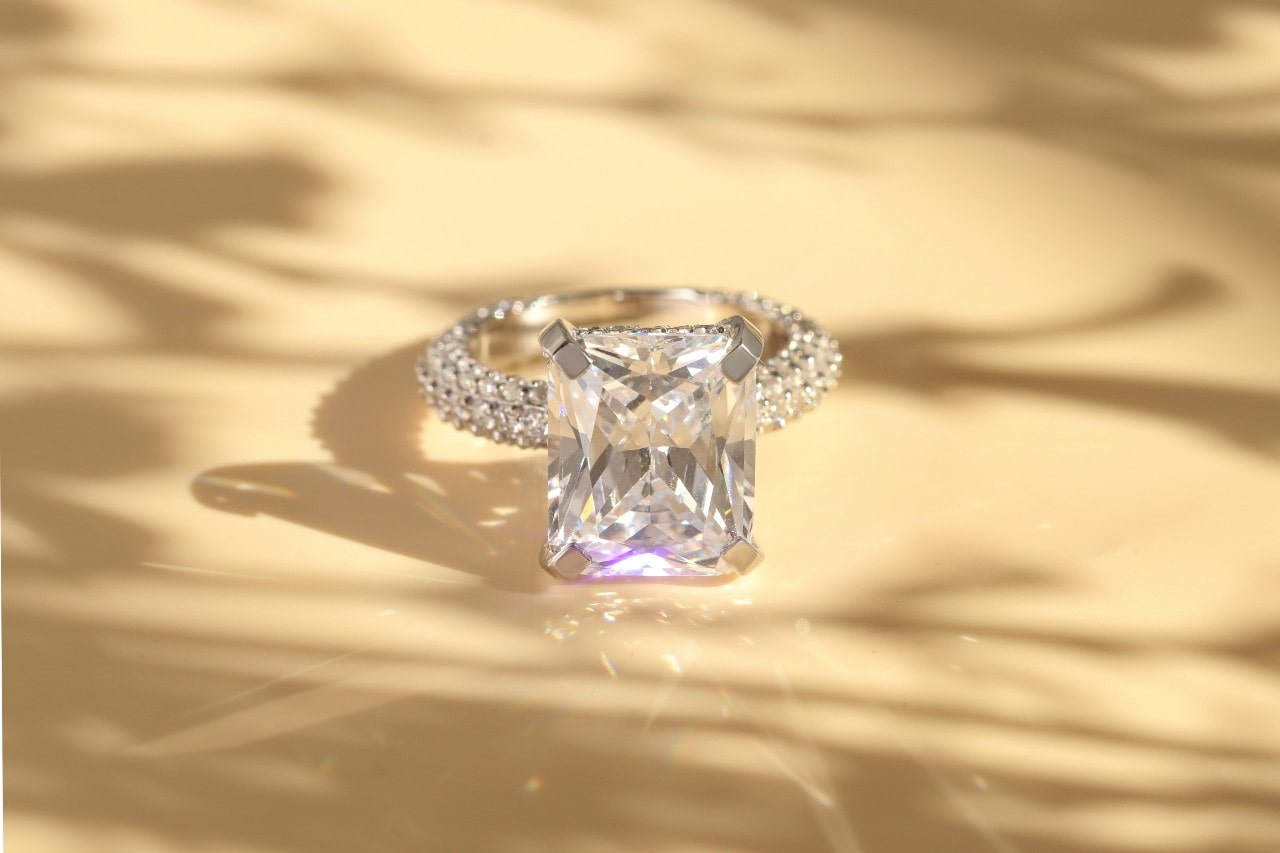 A radiant-cut diamond engagement ring with a pavé band is displayed on a soft, sunlit beige surface with shadow play.