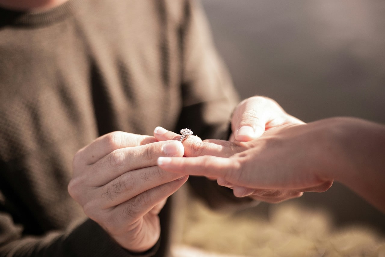 A close up of a man putting a halo engagement ring on a woman’s hand in the outdoor setting.