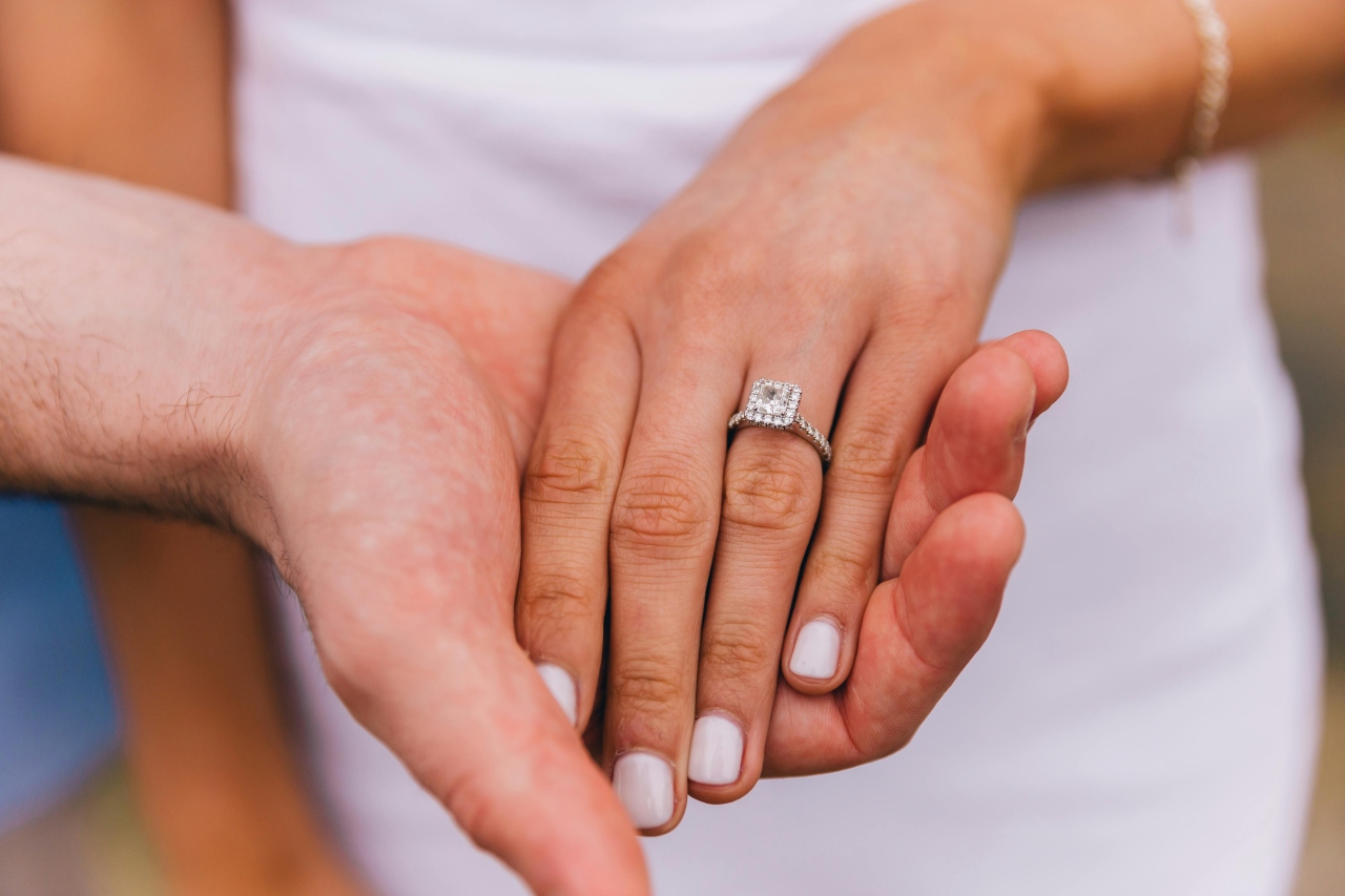 A close of up of a bride’s hand on top of a groom’s hand, showcasing halo white gold diamond engagement ring against a blurry background of her white dress. 