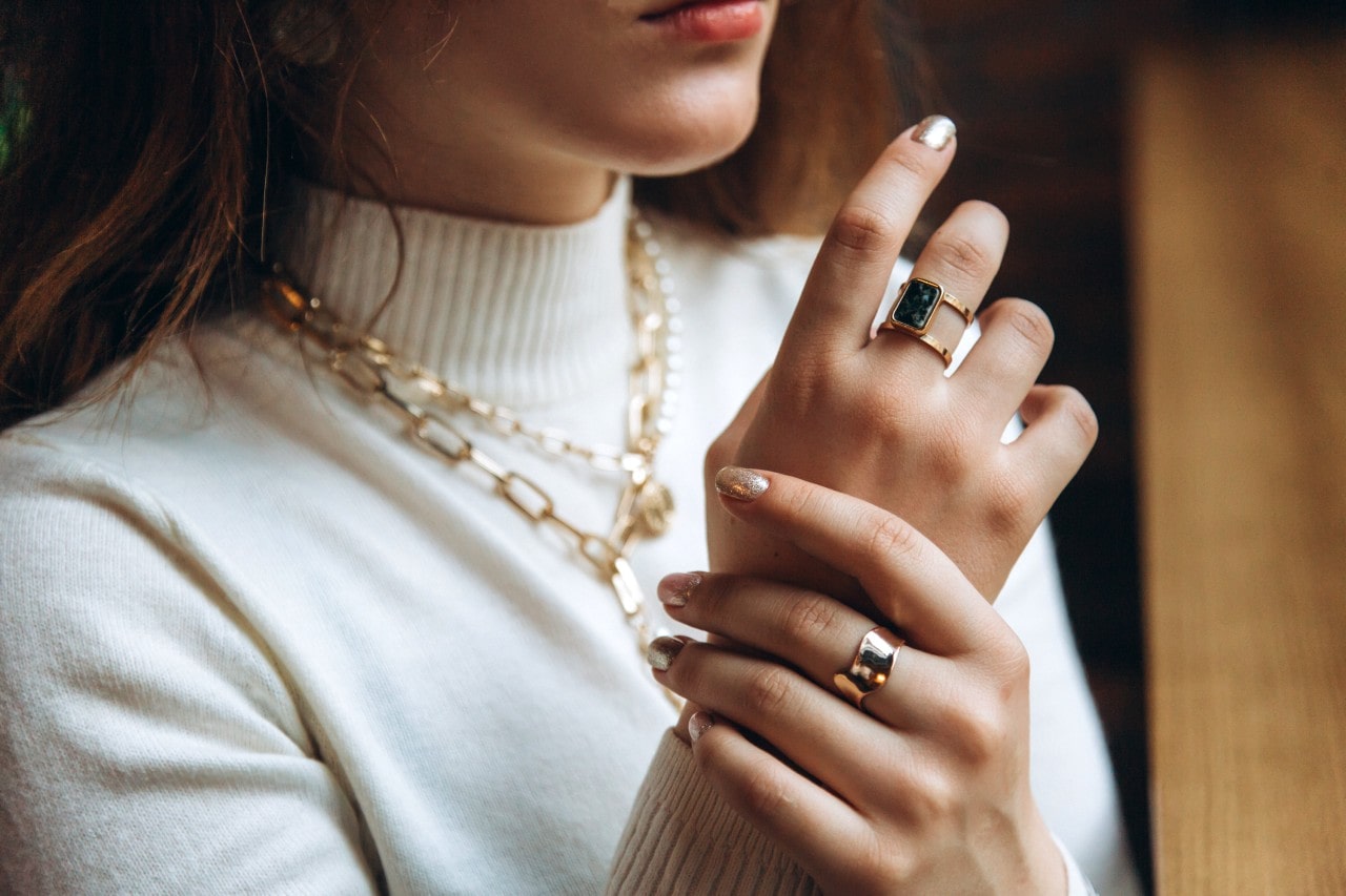 A close up of a woman in white turtleneck, showcasing chunky yellow gold link layered necklace and statement rings.