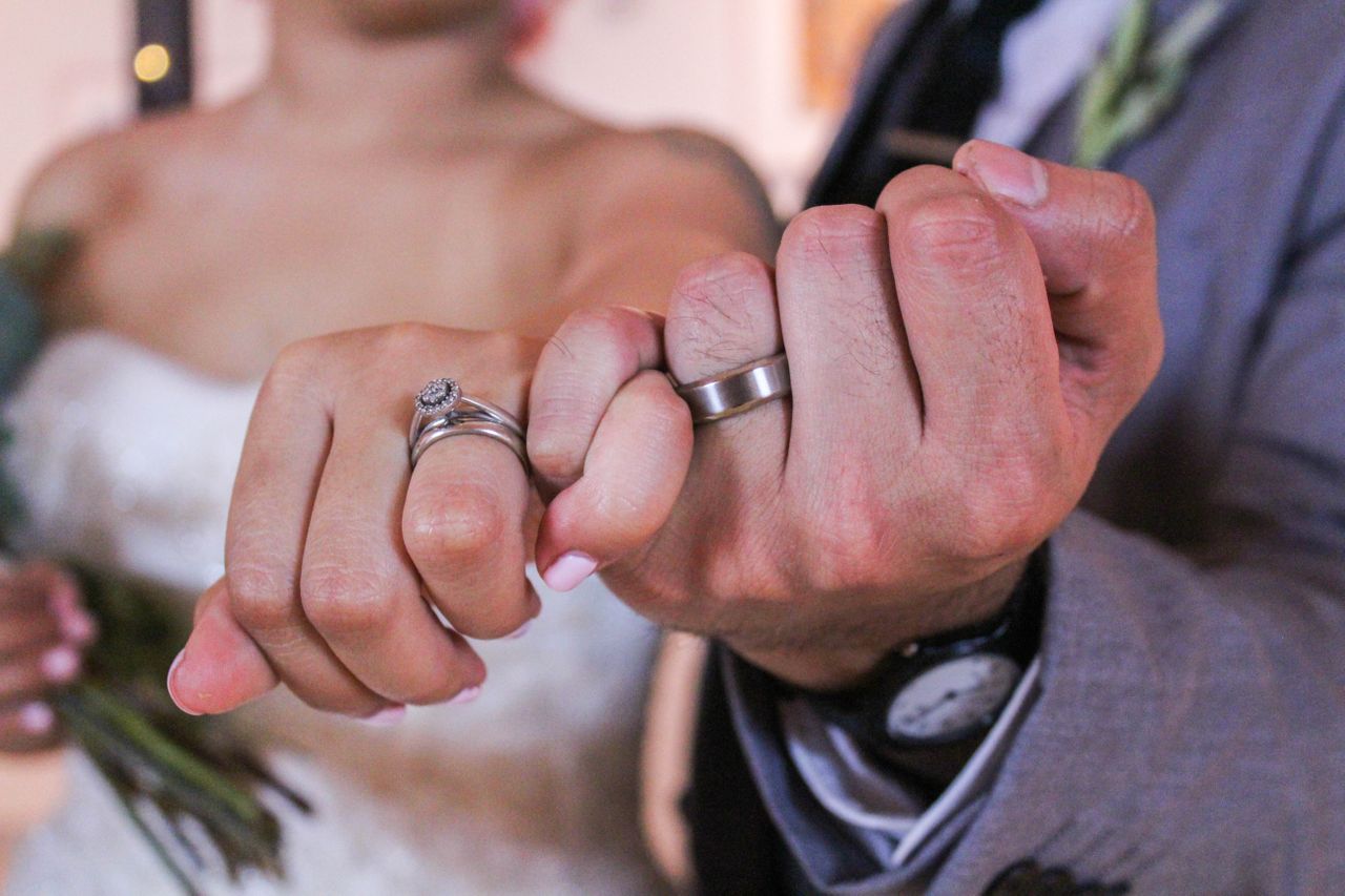 A close-up of newlyweds holding hands, showcasing their rose gold diamond engagement ring and matte wedding band.