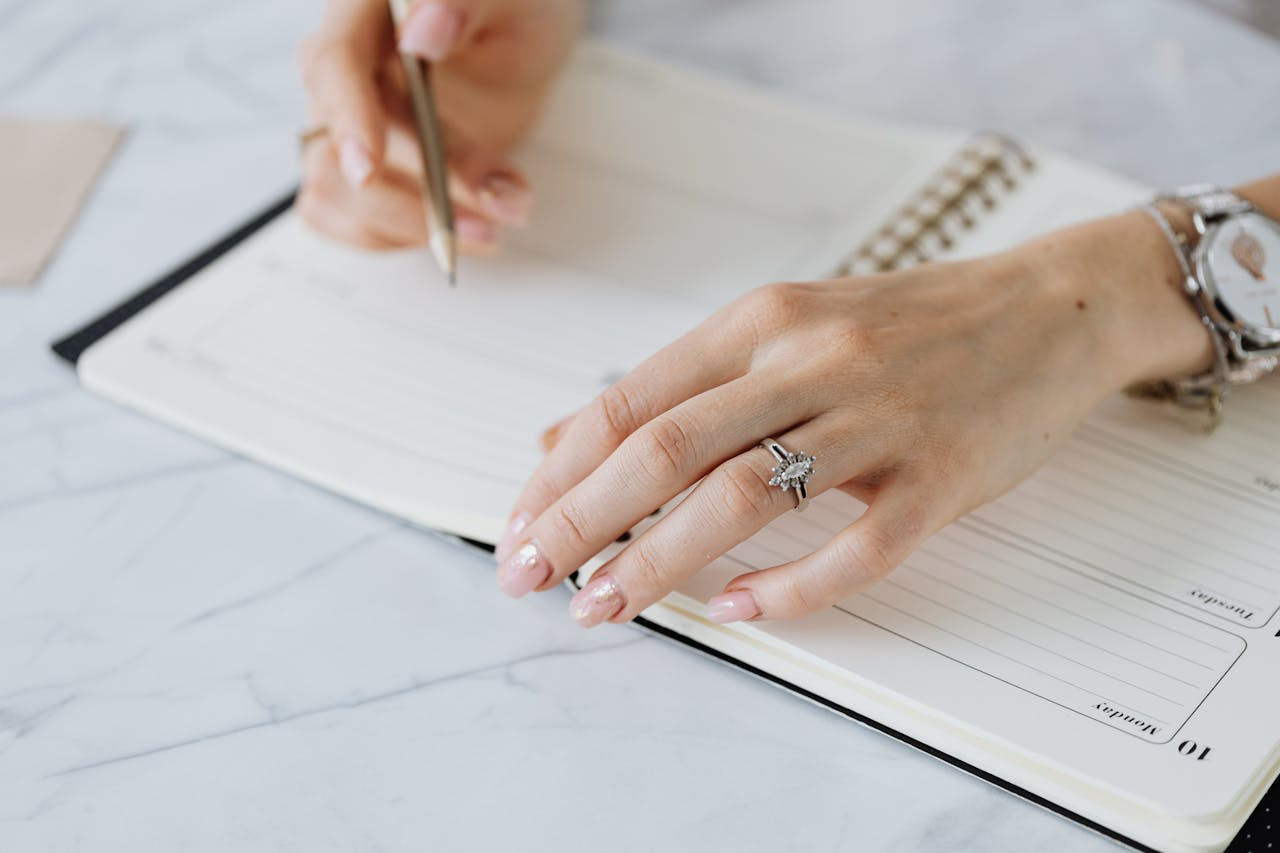 A close up of a woman&rsquo;s hands with long manicured nails writing with a pencil in a notebook, showcasing a marquise cut engagement ring.