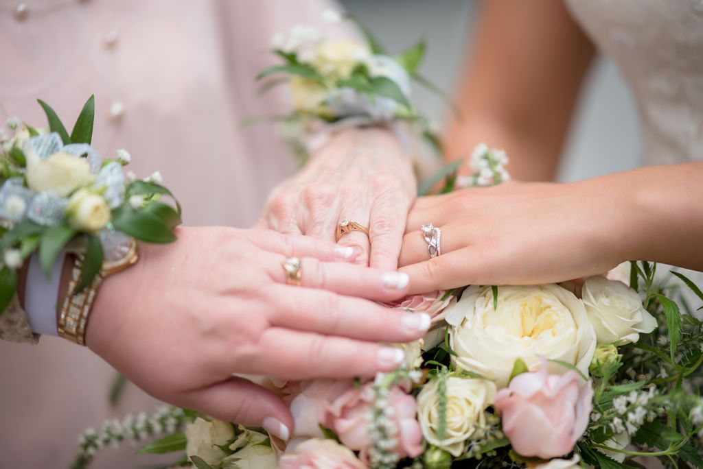 A close-up of three hands of different ages adorned with rings and floral bracelets, resting atop a bouquet of white and pink roses.