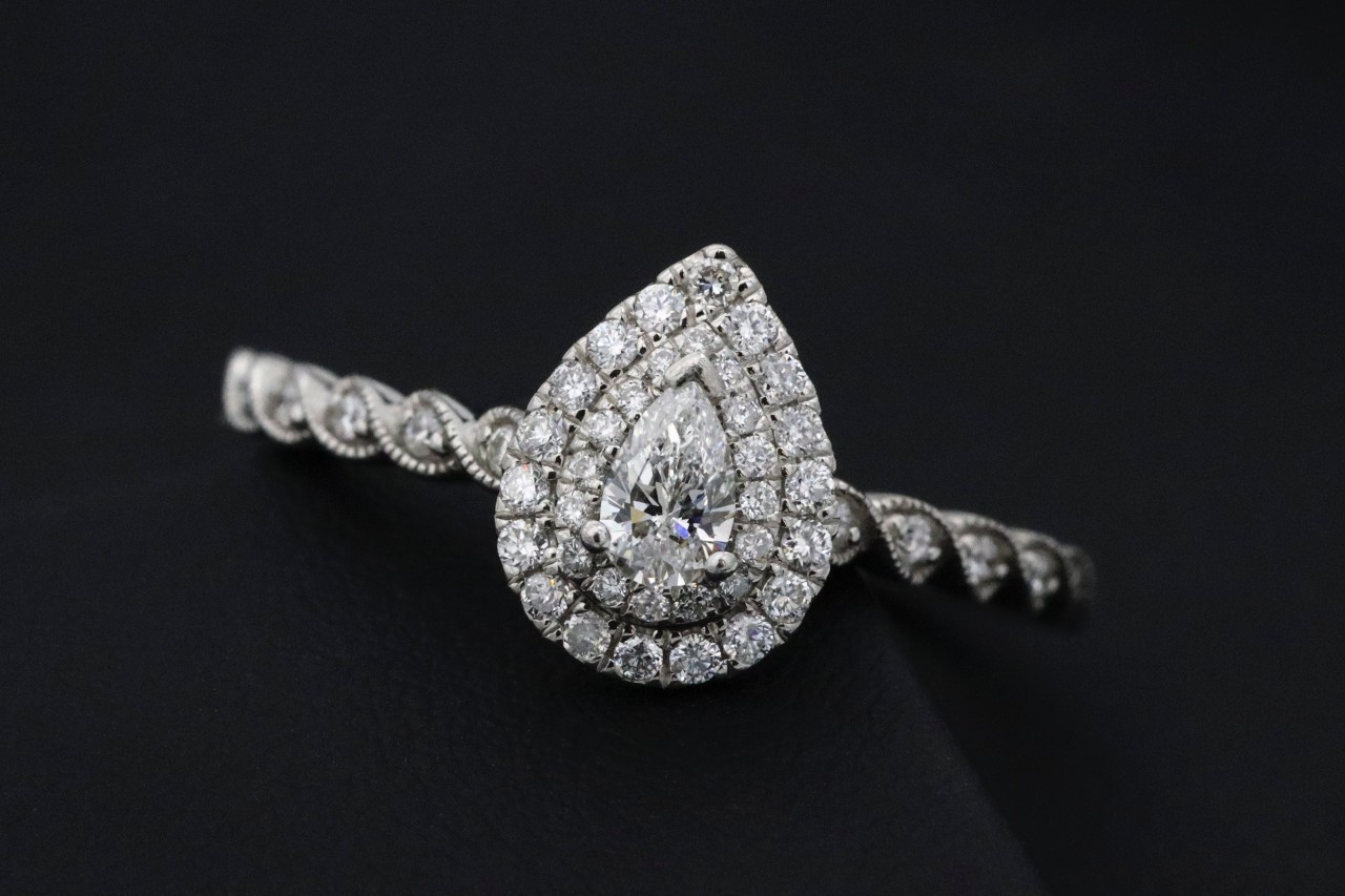 An elegant pear-shaped diamond ring on black background, featuring a central stone with a halo of smaller diamonds and a detailed, twisting band.