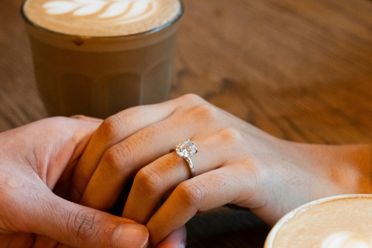 Two hands on a wooden table near the cup of coffee, showcasing oval cut solitaire engagement ring