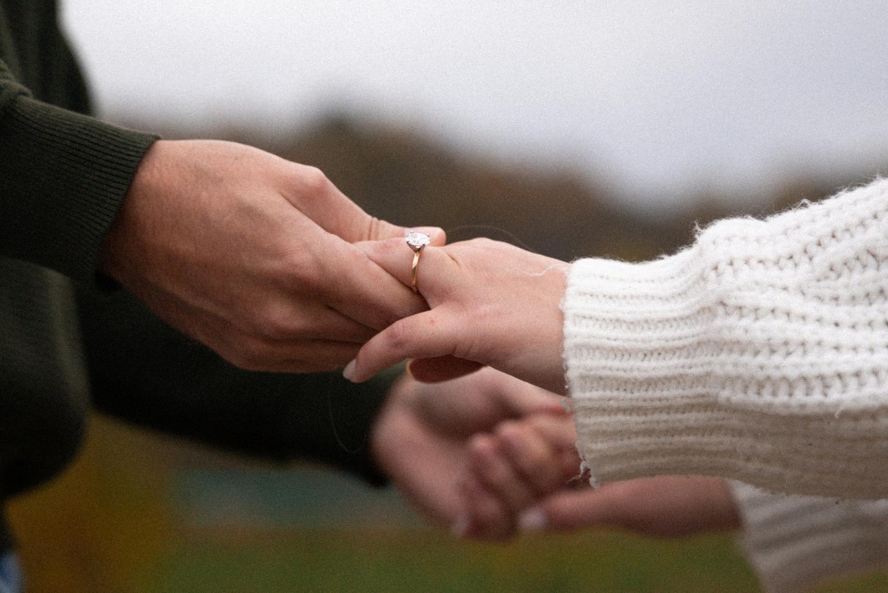 A close up of two intertwined hands, one in white knitted sweater and the other in olive sweatshirt, showcasing yellow gold solitaire engagement rings