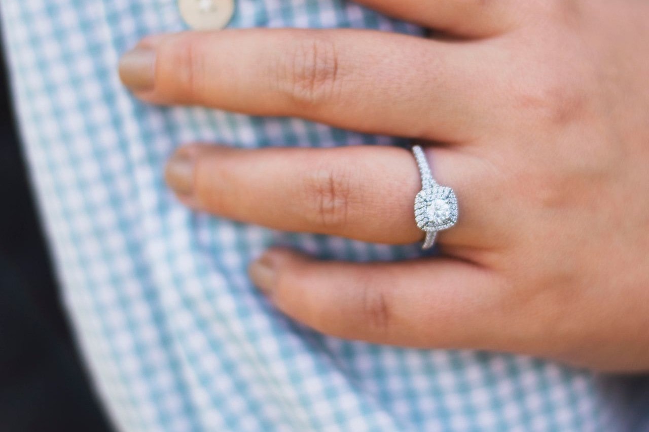 A close-up of a hand on top of the checkered blue shirt adorned with white gold diamond halo engagement ring.