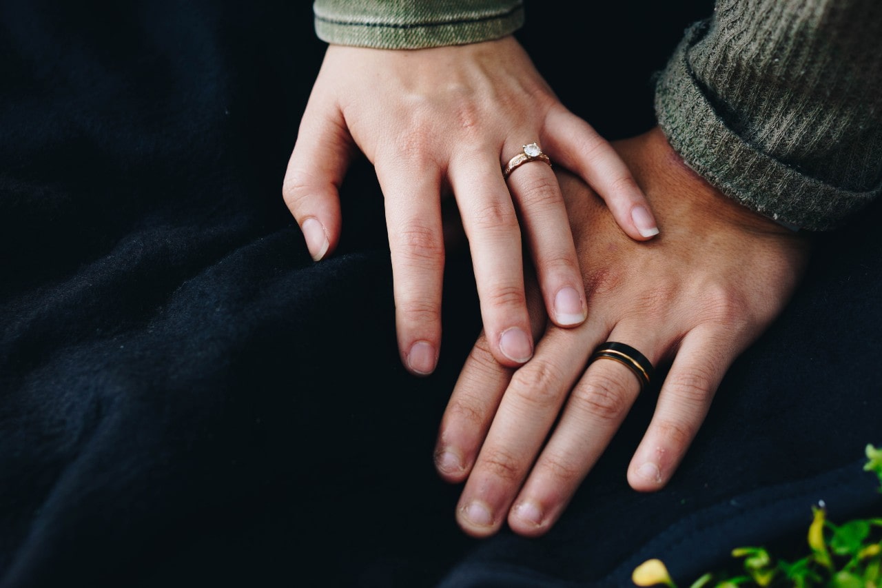 A close-up of a couple’s hand on a dark blue blanket, showcasing men’s mixed metal wedding band and woman’s rose gold diamond engagement ring. A close-up of a couple’s hand on a dark blue blanket, showcasing men’s mixed metal wedding band and woman’s rose gold diamond engagement ring.