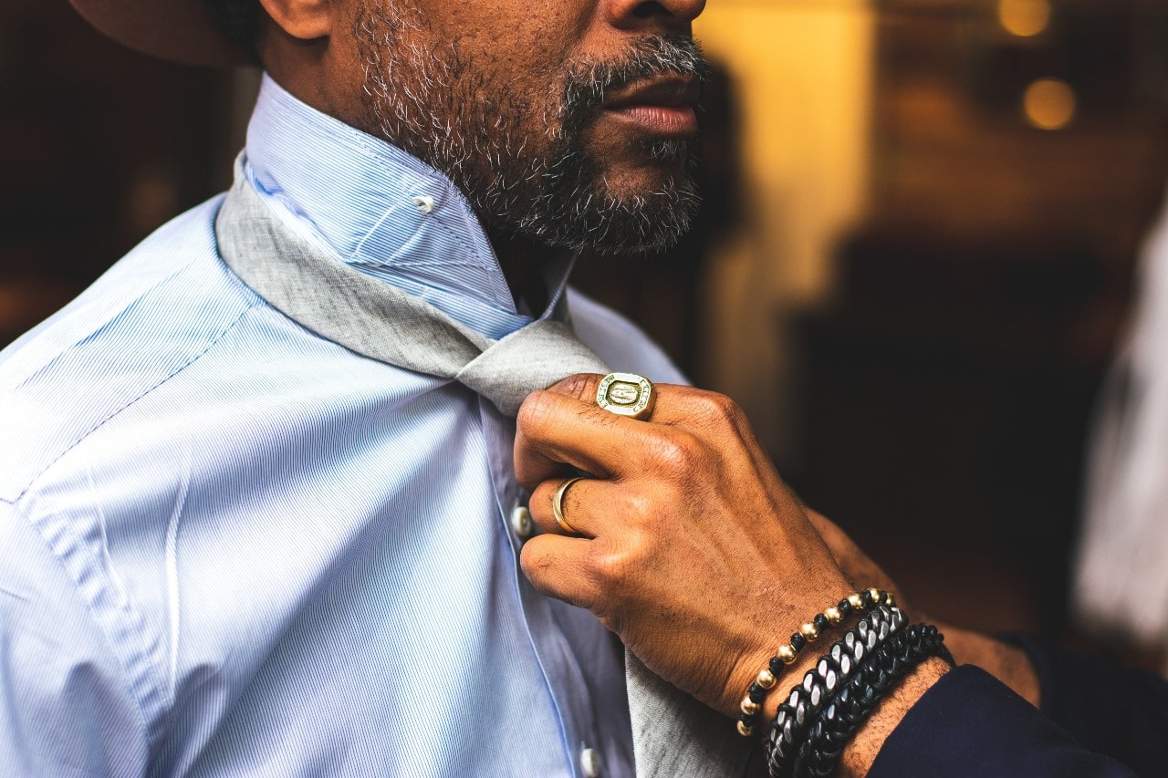 A man with a beard wears rings and bracelets while another person adjusts his light gray tie. A man with a beard wears rings and bracelets while another person adjusts his light gray tie.