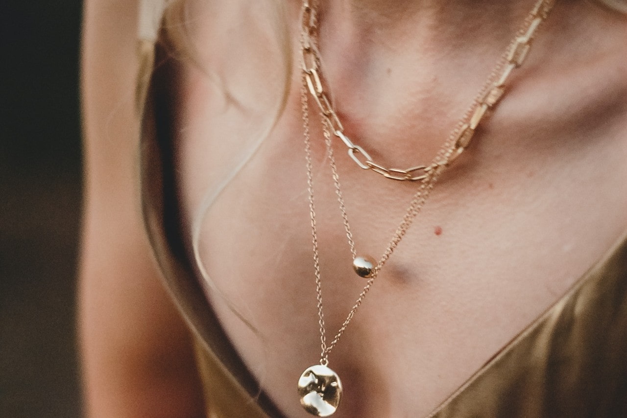 Close-up of a woman wearing layered gold necklaces; one with a chain-link design, the other with small gold pendants, against a satin garment.
