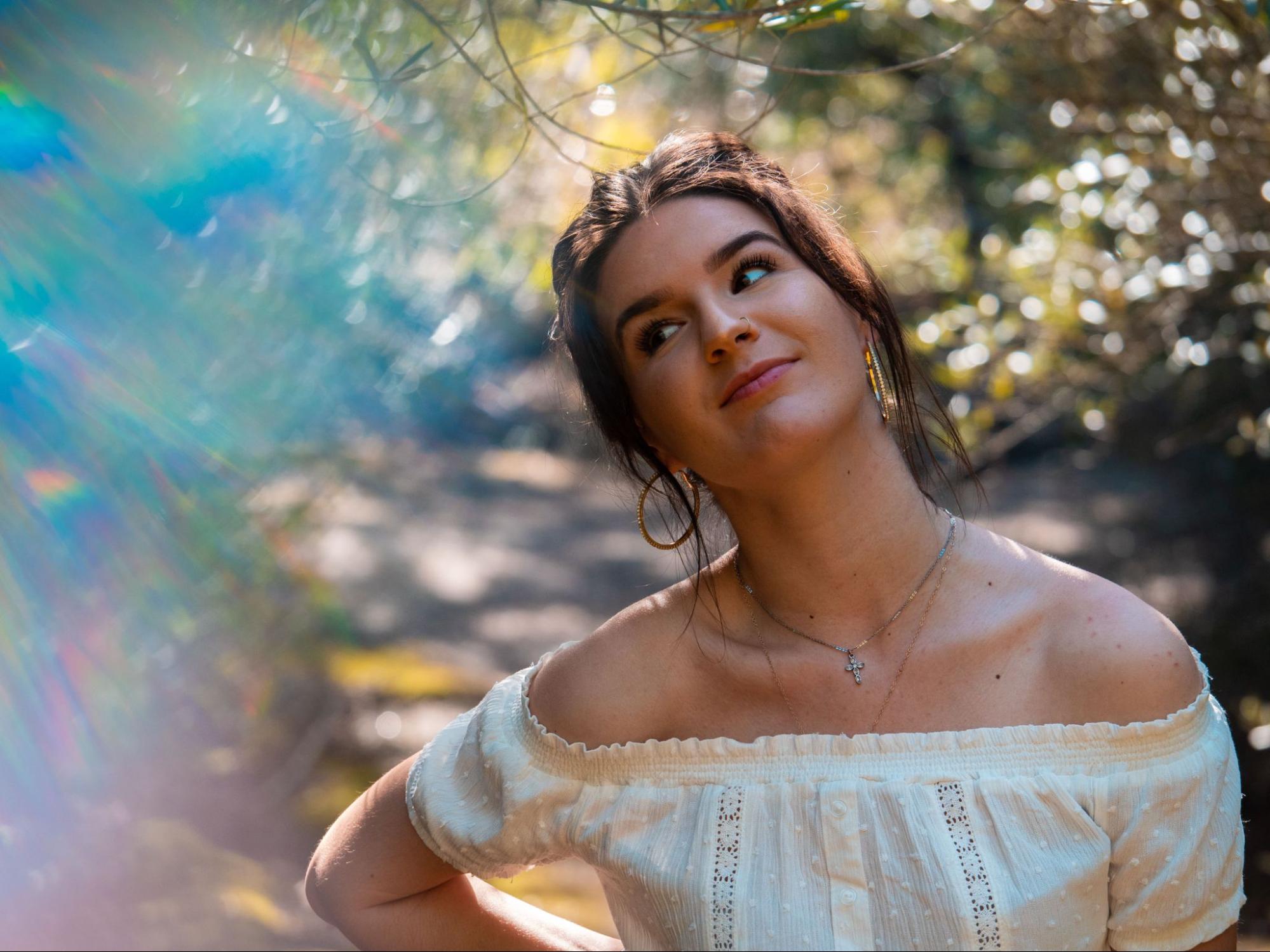 Young woman in an off-the-shoulder white blouse looks upward with a thoughtful expression, showcasing a silver cross necklace.
