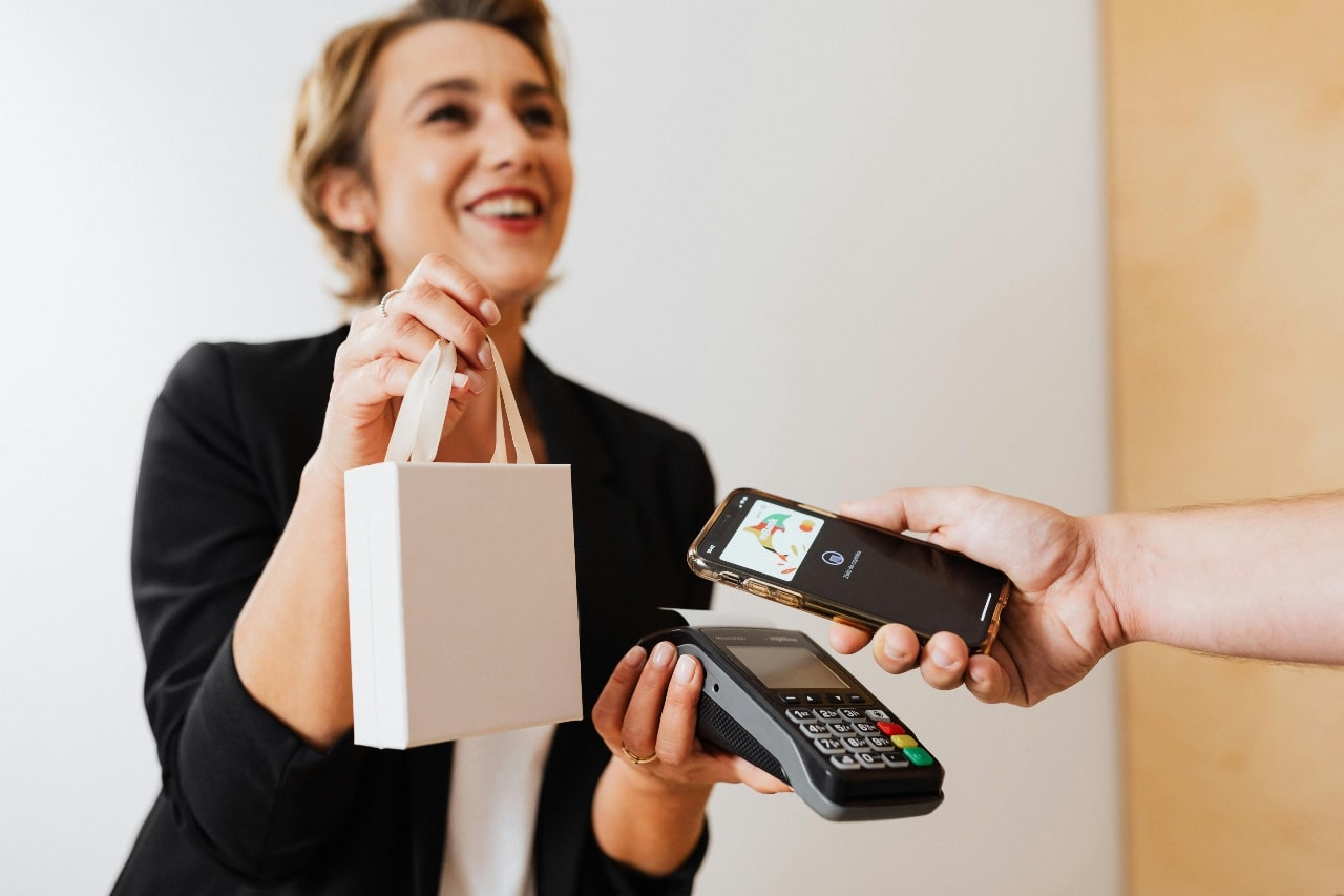 Smiling woman holding a pink shopping bag while a person makes a contactless payment with a smartphone on a card reader.