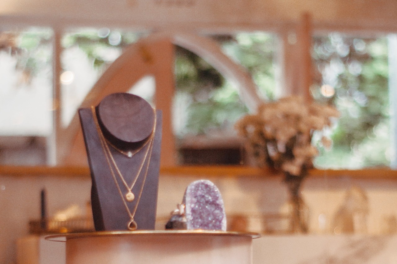 A display of layered gold necklaces on a black bust in a softly lit jewelry store, with a bouquet of flowers and a purple geode in the background.