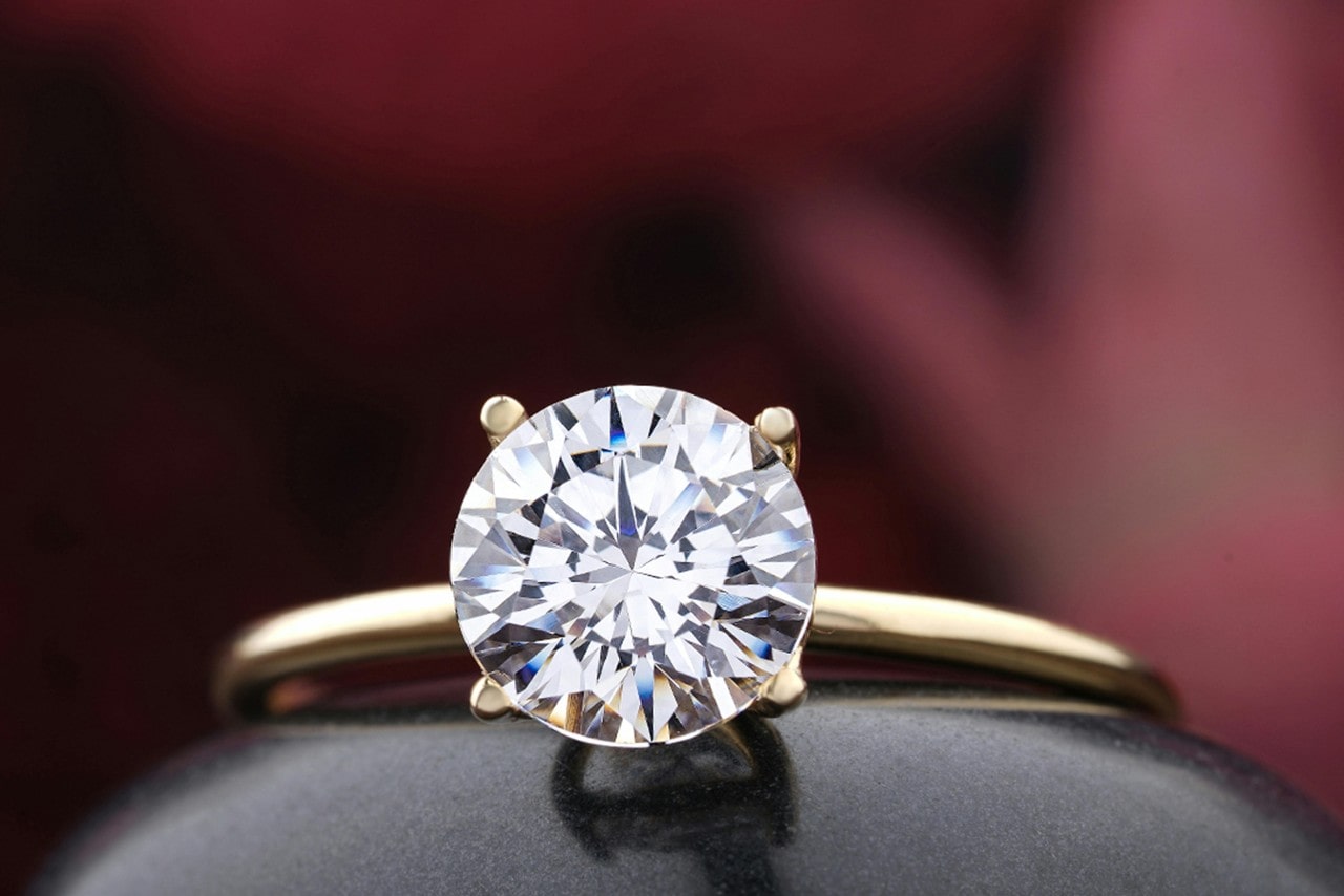 A yellow gold solitaire round cut diamond engagement ring in front of the burgundy blurry background