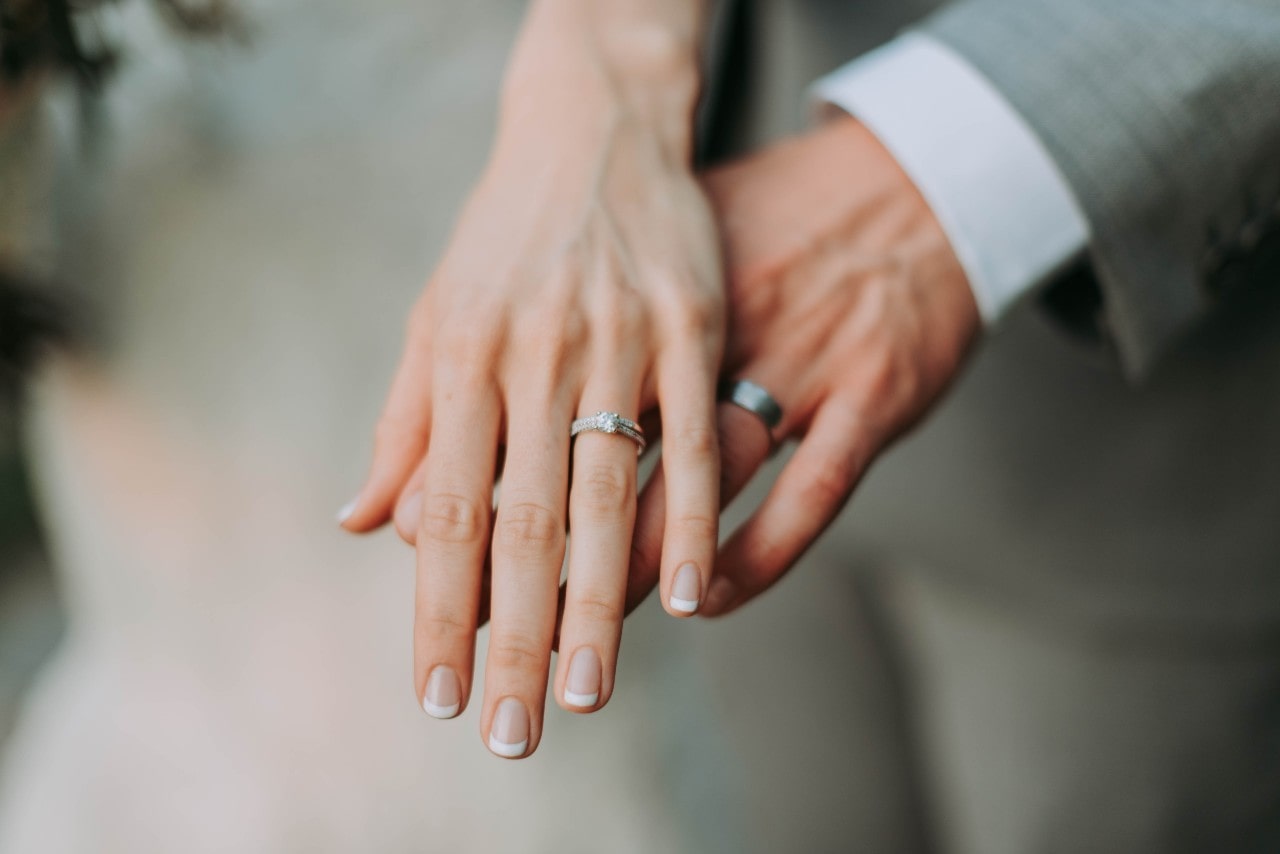A close up of two hands showing matching wedding bands, with the bride’s diamond ring and French-manicured nails in focus.