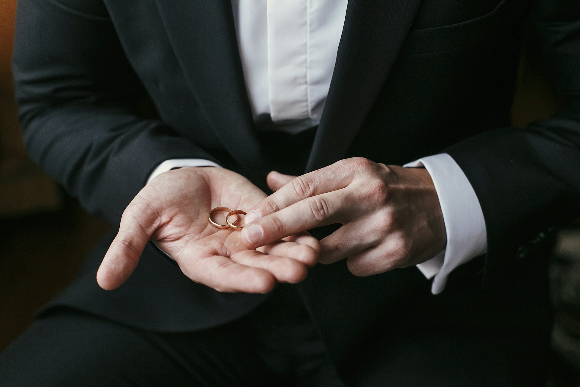 A close up of a man in a black suit holding two thing yellow golf wedding bands in his palm.