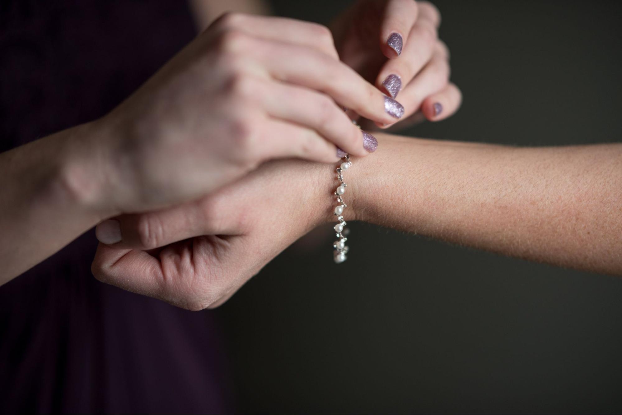 A person is fastening a delicate pearl and crystal bracelet onto another person’s wrist, both with well-manicured hands.