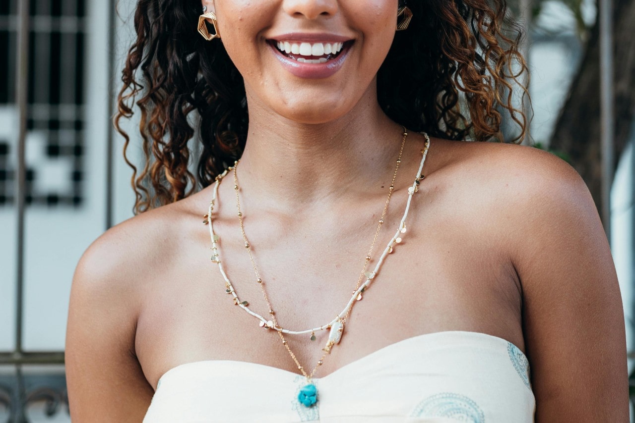 A close-up of a woman smiling in a white strapless top, showcasing a layered yellow gold necklace with a blue stone in the center.