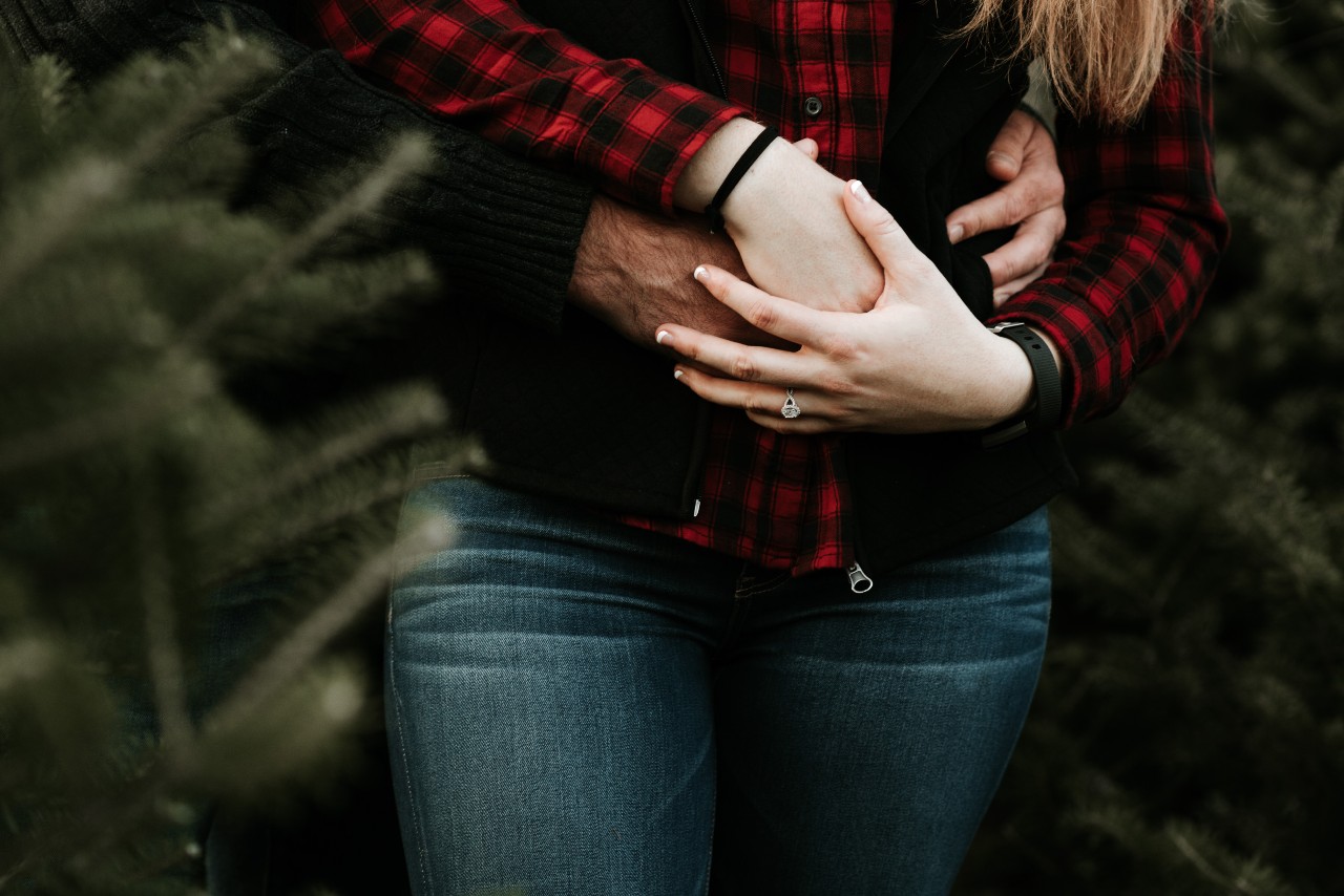A couple standing together in pine trees, the halo engagement ring on her hand is visible as the man hugs her and has his arms around her waist A couple standing together in pine trees, the halo engagement ring on her hand is visible as the man hugs her and has his arms around her waist
