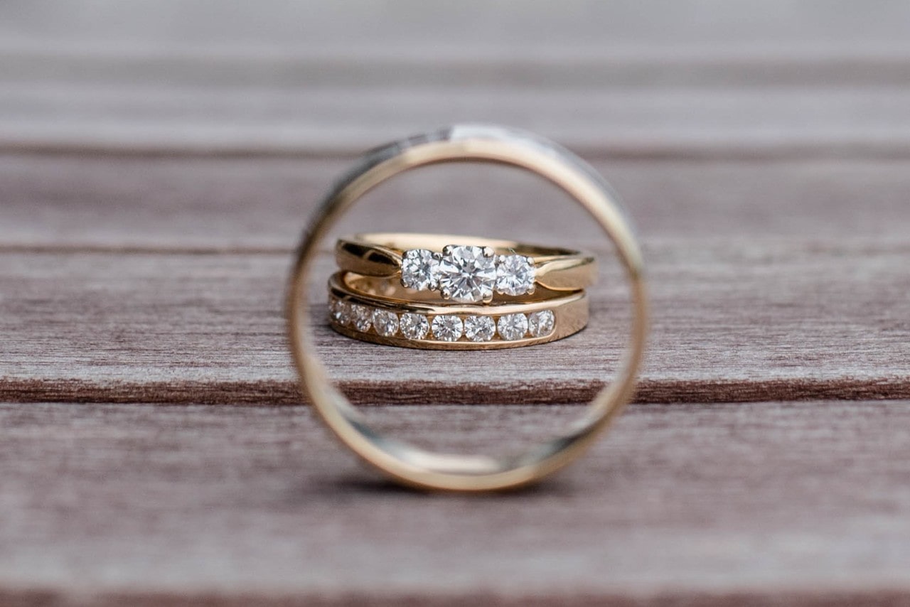 A yellow-gold men’s wedding band frames a bride’s wedding stack, with a three stone engagement ring and a diamond band