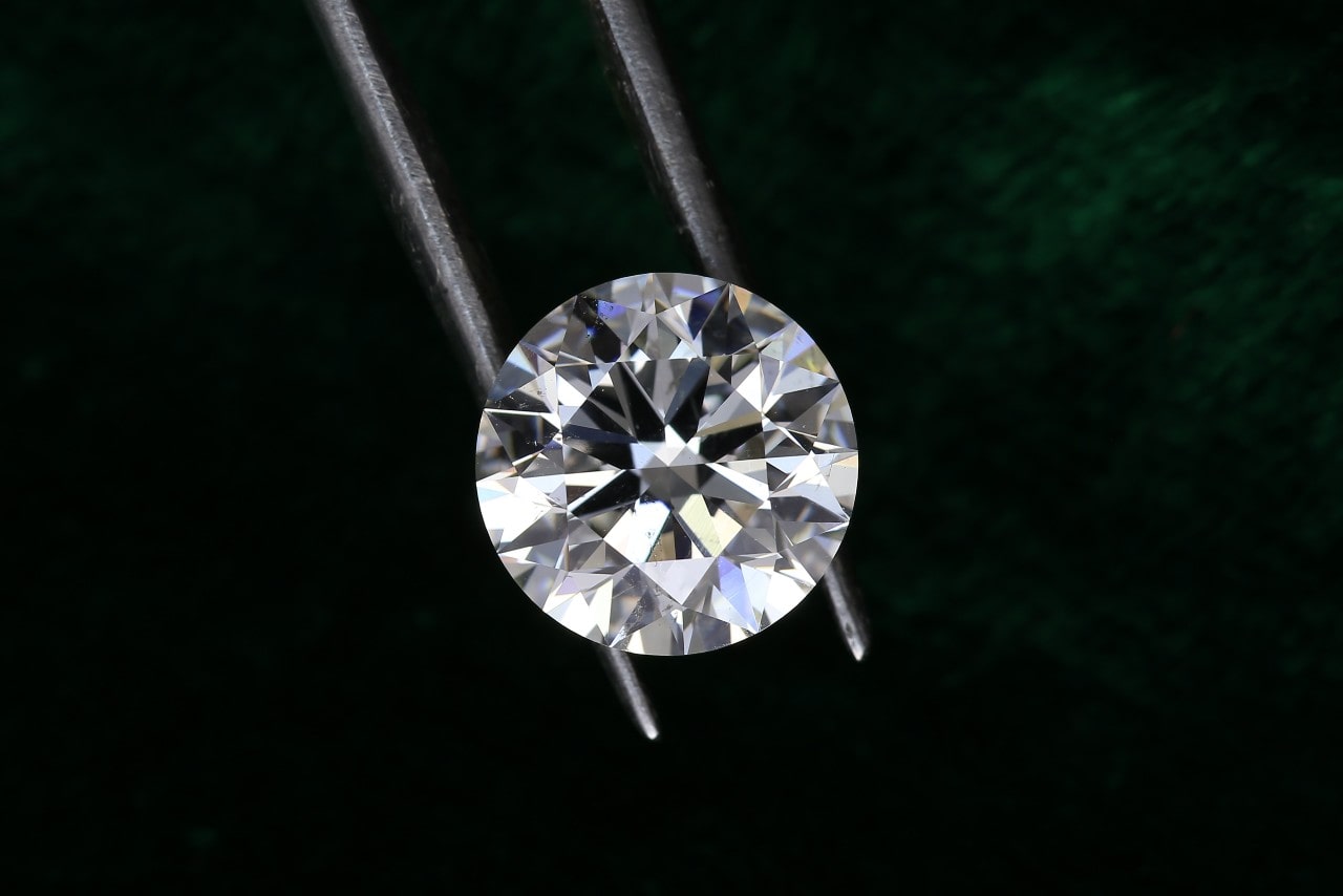 A professional jeweller holds a round-cut lab-created diamond with a pair of jewellery tweezers against a dark green background