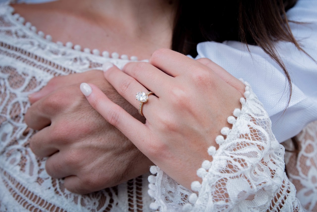 A large oval centre stone with a dainty diamond covered band being worn by a boho bride holds the wrist of her groom who has wrapped their arm around her