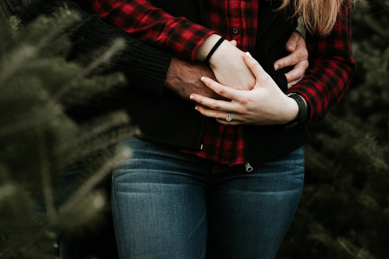 A couple standing together in pine trees, the halo engagement ring on her hand is visible as the man hugs her and has his arms around her waist