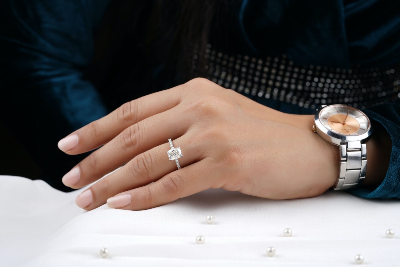 A woman wearing an emerald side stone engagement ring and a silver watch with a blue silk long sleeve shirt with a display of pearls in front of her