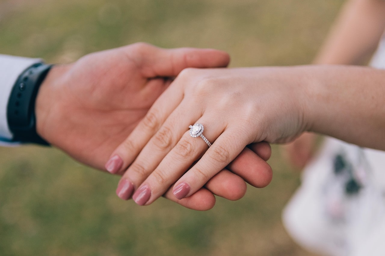 A woman holding the hand of her love while wearing an oval halo engagement ring with diamonds along the band