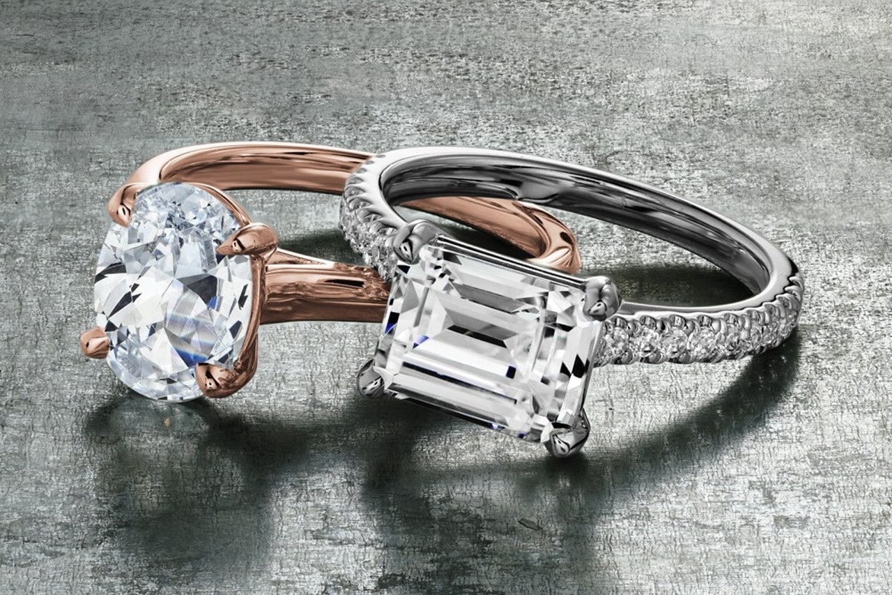 A rose gold, oval cut engagement ring next to a silver, emerald cut engagement ring