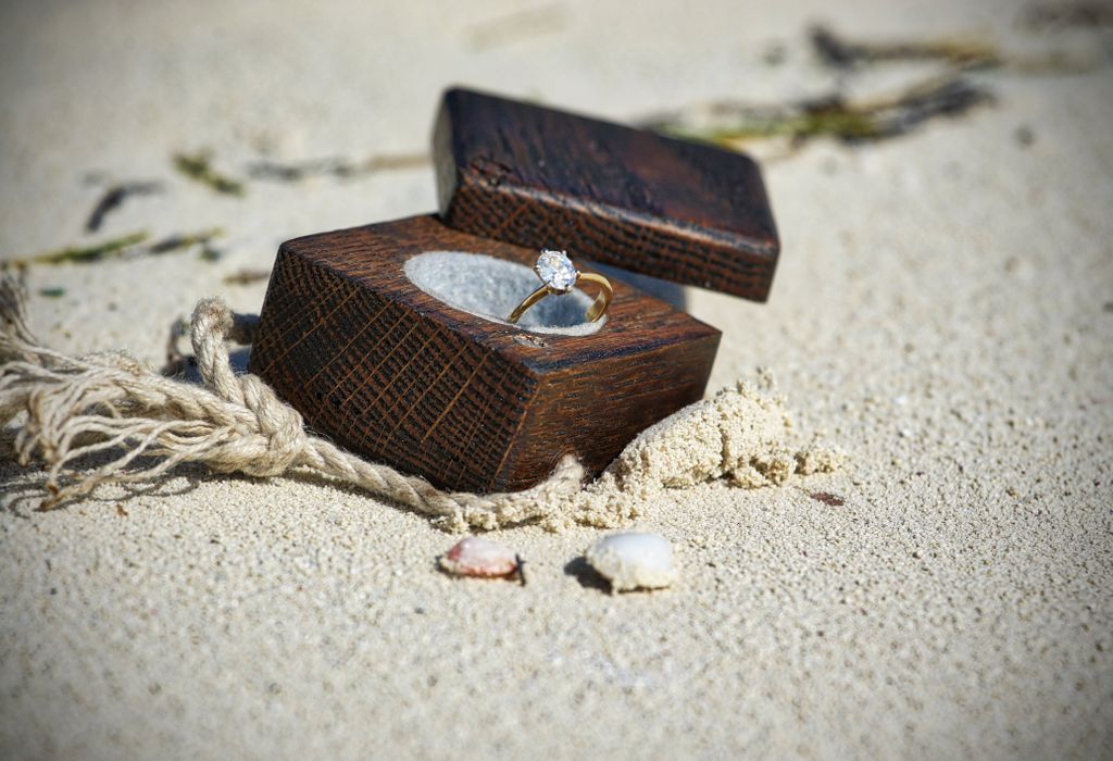 A gold oval-cut engagement ring sits in a wooden box in the sand