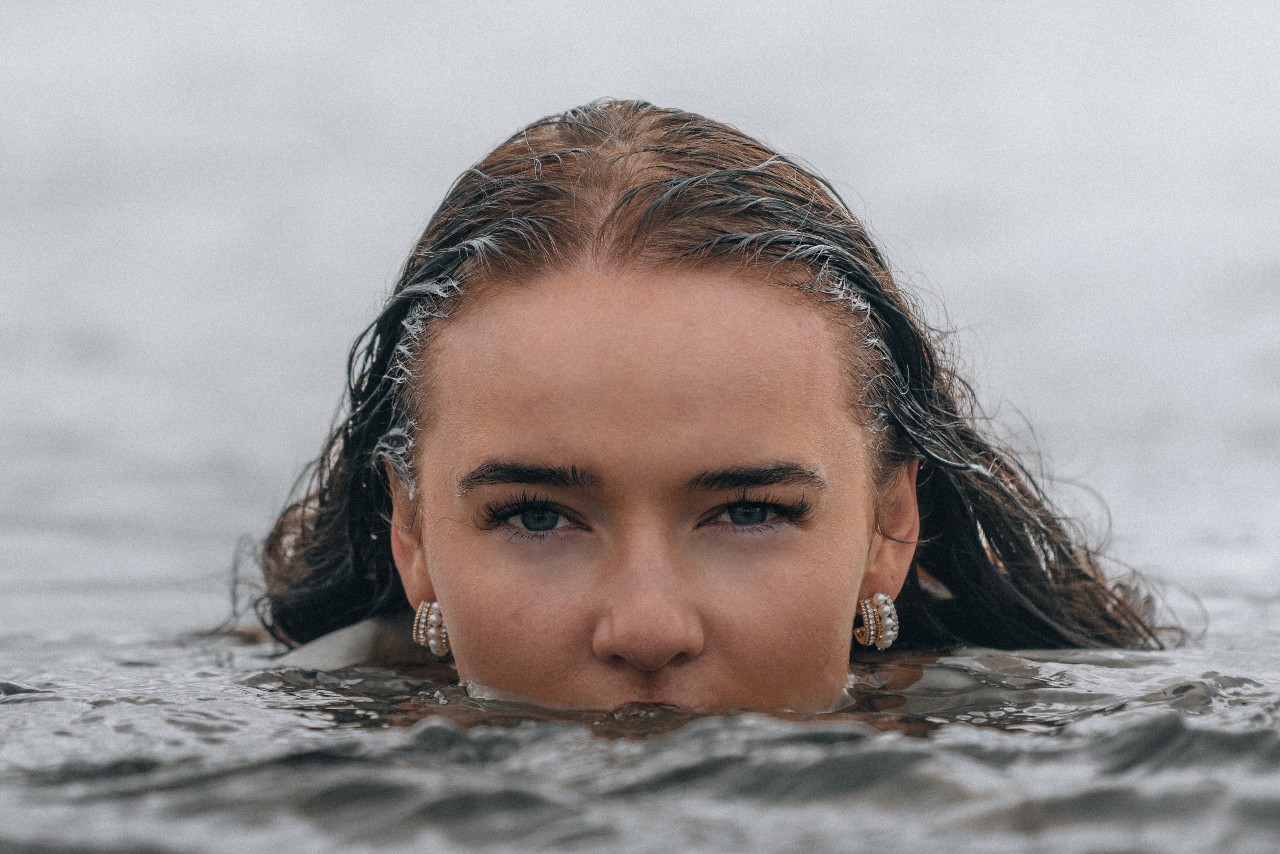 a woman whose face is half submerged in water, wearing pearl huggies earrings