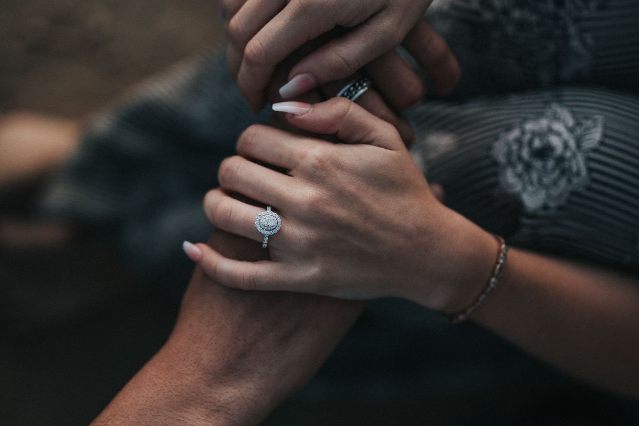 A closeup of a couple joining hands, the woman wearing a halo engagement ring.