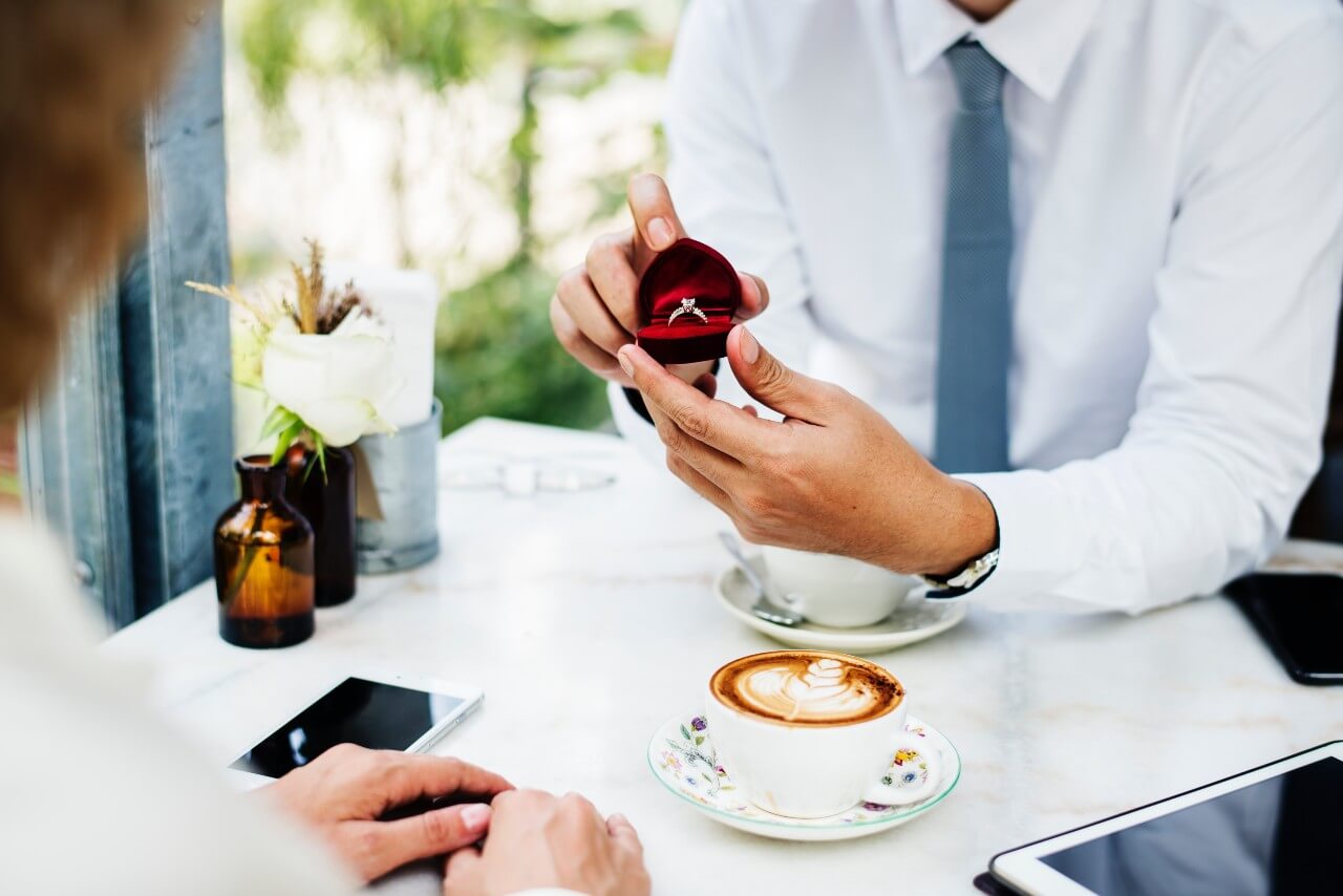 A man opens a ring box and proposes at a cafe table.