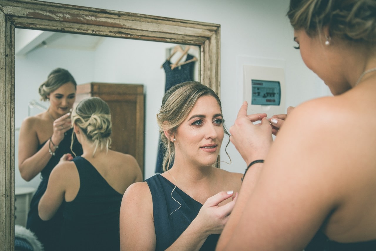 Two bridesmaids working on putting on a necklace in front of a mirror